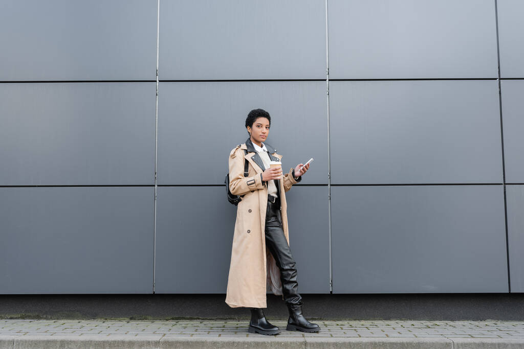 longitud completa de la mujer afroamericana en gabardina beige de pie con bebida para llevar y teléfono inteligente cerca de la pared gris en la calle - Foto, Imagen