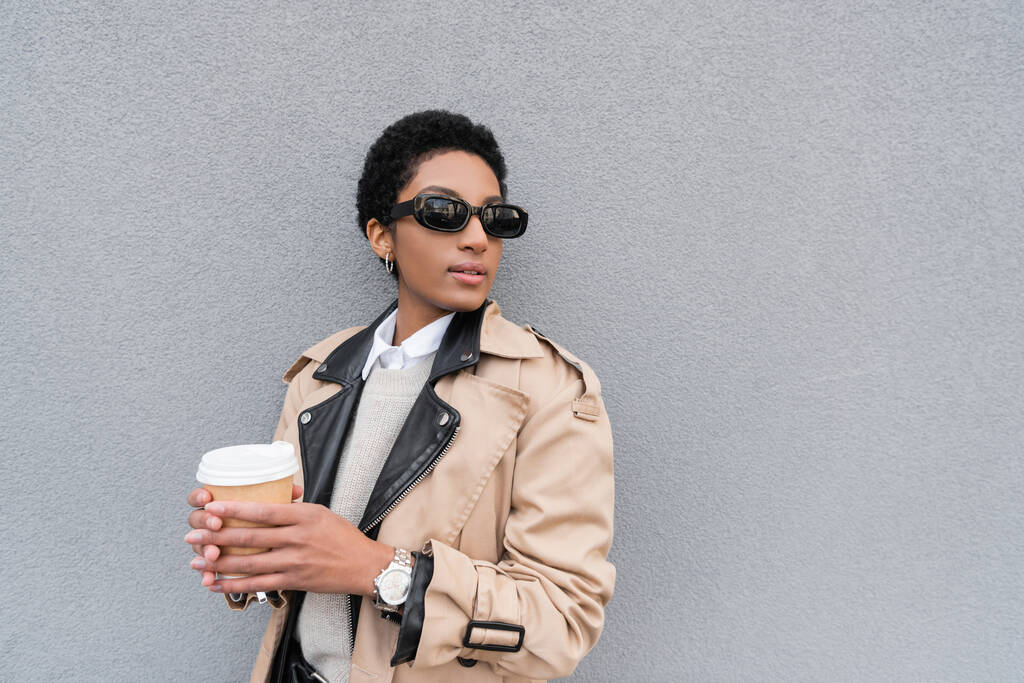 mujer de negocios afroamericana en gabardina beige y gafas de sol sosteniendo taza de papel y mirando hacia otro lado cerca de la pared gris en la ciudad - Foto, Imagen