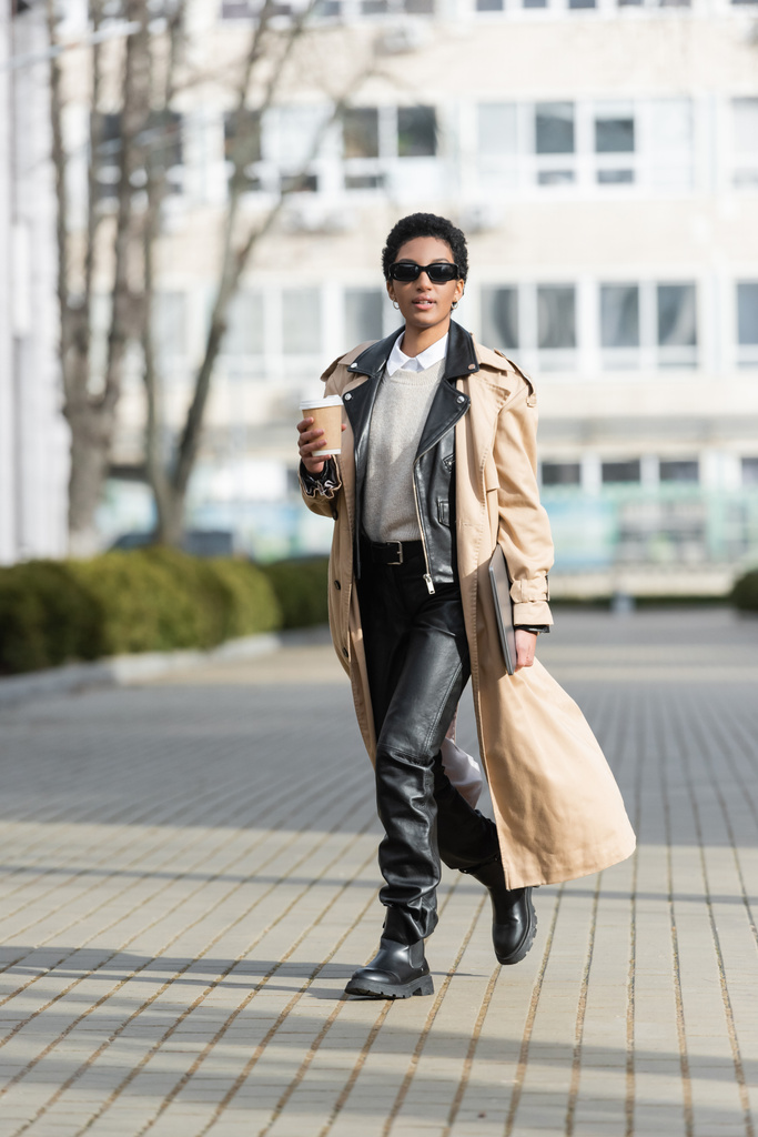 Siyah deri pantolonlu, trençkotlu, laptoplu ve kağıt bardaklı Afro-Amerikan iş kadını. Bulanık sokakta yürürken. - Fotoğraf, Görsel