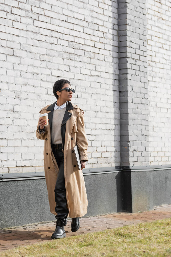 volledige lengte van Afrikaanse Amerikaanse zakenvrouw in zonnebril en trench jas lopen met laptop en koffie te gaan in de buurt van grijze muur op stedelijke straat - Foto, afbeelding