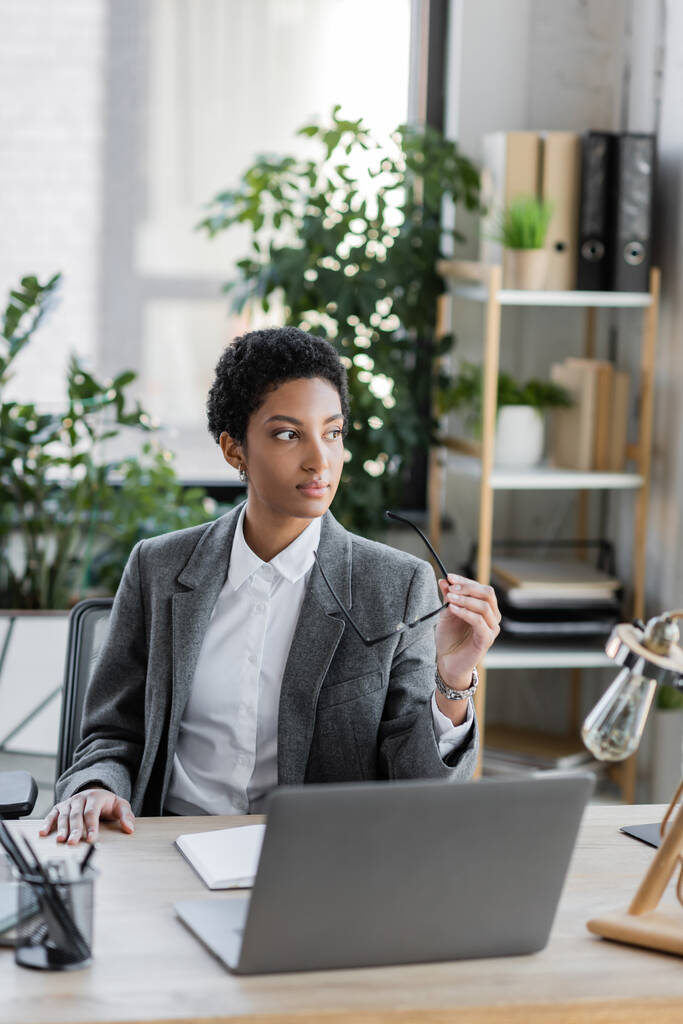 στοχαστική αφροαμερικανίδα επιχειρηματίας με κοστούμι κρατώντας γυαλιά ενώ κάθεται στο χώρο εργασίας στο γραφείο - Φωτογραφία, εικόνα