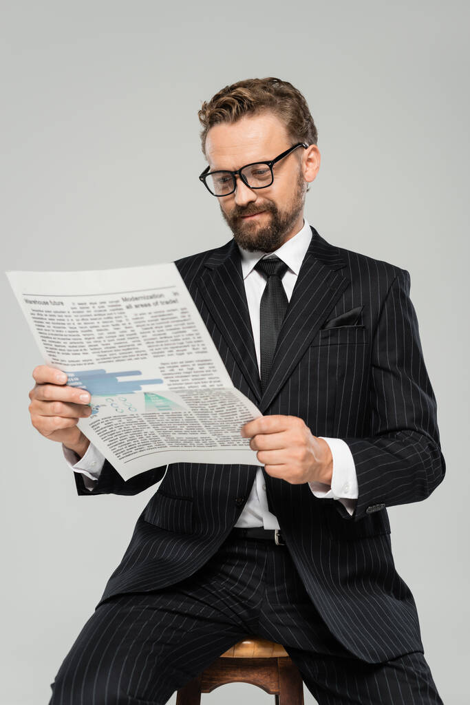 empresario complacido en traje y gafas leyendo periódico aislado en gris  - Foto, imagen