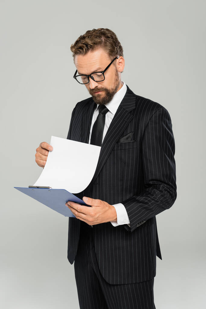 hombre de negocios bien vestido con gafas sujetando portapapeles y papel aislado en gris  - Foto, imagen