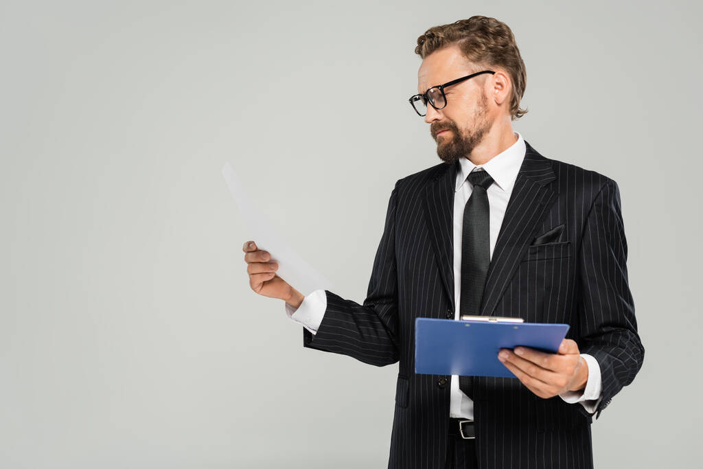 hombre de negocios bien vestido con gafas sujetando el portapapeles y mirando el papel aislado en gris  - Foto, Imagen
