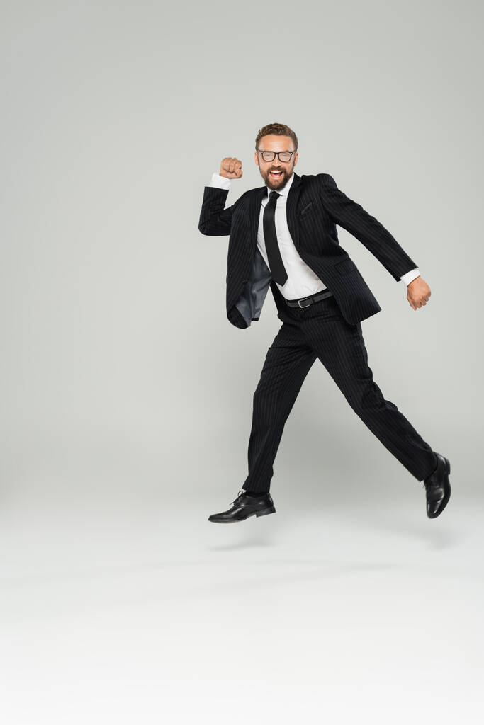 full length of excited businessman in glasses and suit jumping on grey  - Photo, Image