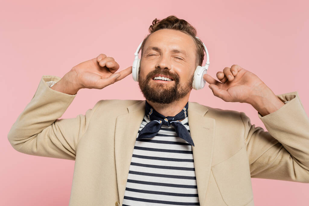 満足したフランス人男性でブレザーと首のスカーフで聴く音楽でワイヤレスヘッドフォンでピンクに隔離  - 写真・画像