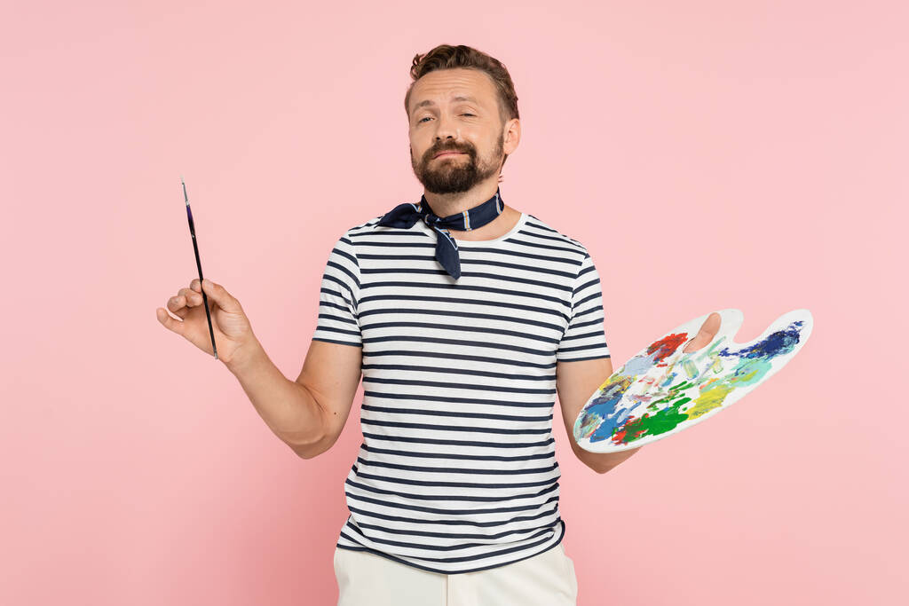 bearded french artist with neck scarf holding palette with colorful paints and paintbrush isolated on pink - Photo, Image