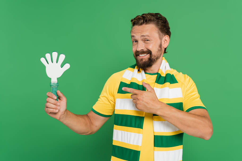 torcedor de futebol alegre em cachecol listrado apontando com o dedo em palmas de mão de plástico isolado em verde  - Foto, Imagem