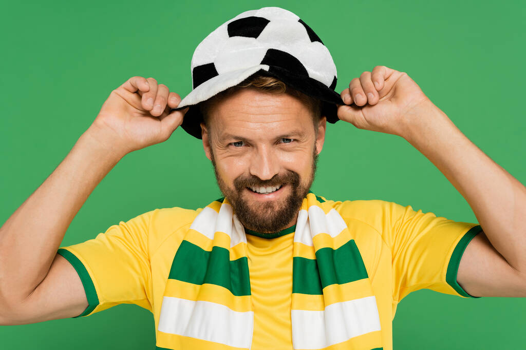 счастливый бородатый мужчина в полосатом шарфе в футбольной шляпе фаната изолированы на зеленый  - Фото, изображение