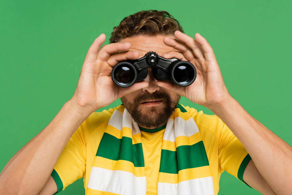 homme barbu en écharpe rayée regardant à travers des jumelles pendant le match de football isolé sur vert - Photo, image