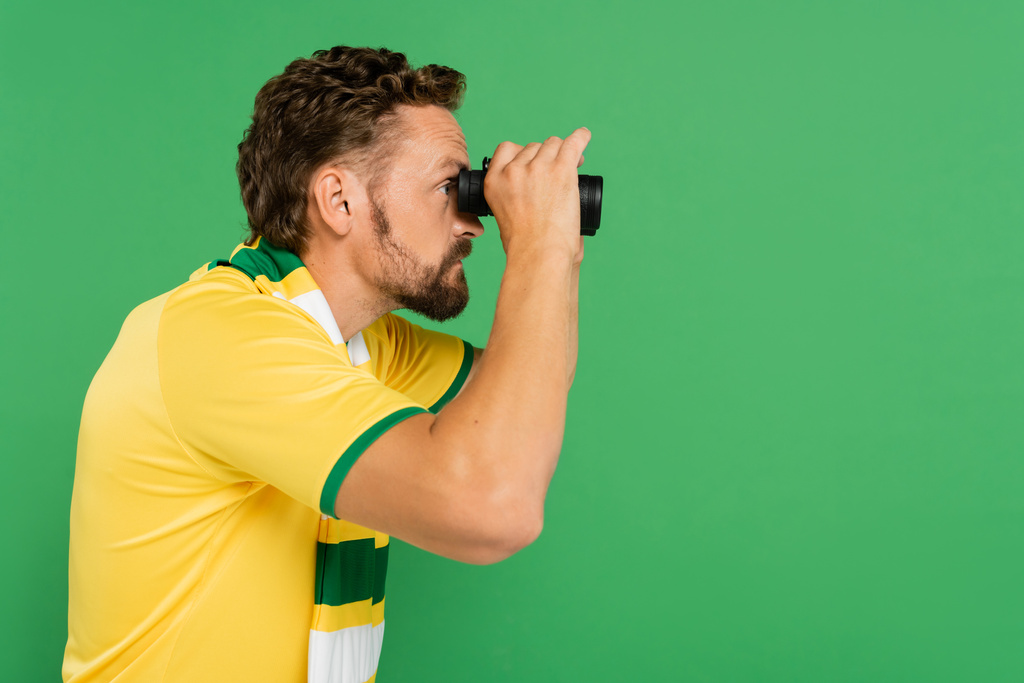 visão lateral do homem barbudo em lenço listrado segurando binóculos durante jogo de futebol isolado em verde - Foto, Imagem