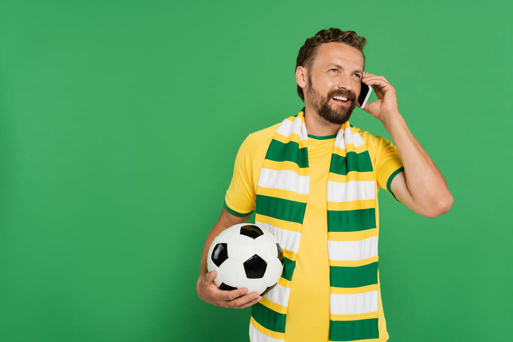 Χαρούμενος άντρας με ριγέ φουλάρι που κρατάει το ποδόσφαιρο και μιλάει στο κινητό τηλέφωνο απομονωμένος στο πράσινο - Φωτογραφία, εικόνα