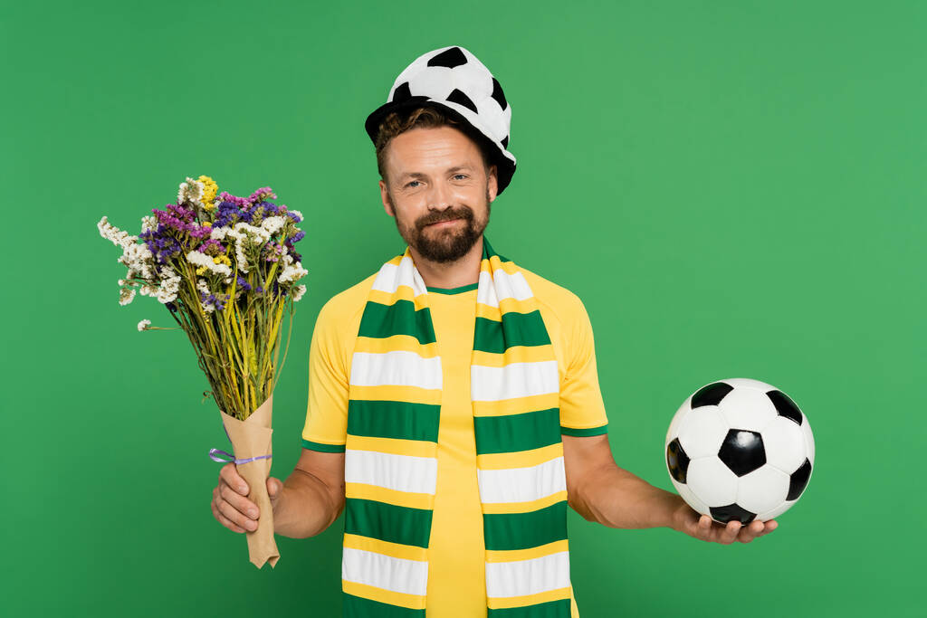 uomo barbuto in cappello tifoso sport in possesso di calcio e fiori selvatici isolati su verde  - Foto, immagini