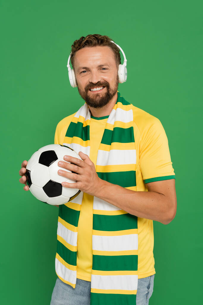 feliz fã de esportes barbudos em cachecol e fones de ouvido segurando futebol isolado em verde - Foto, Imagem