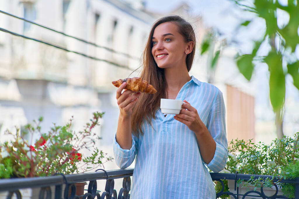 Retrato de una hermosa feliz linda alegre mujer romántica sonriente con taza de café y croissant recién horneado para el desayuno francés por la mañana en un balcón                      - Foto, imagen