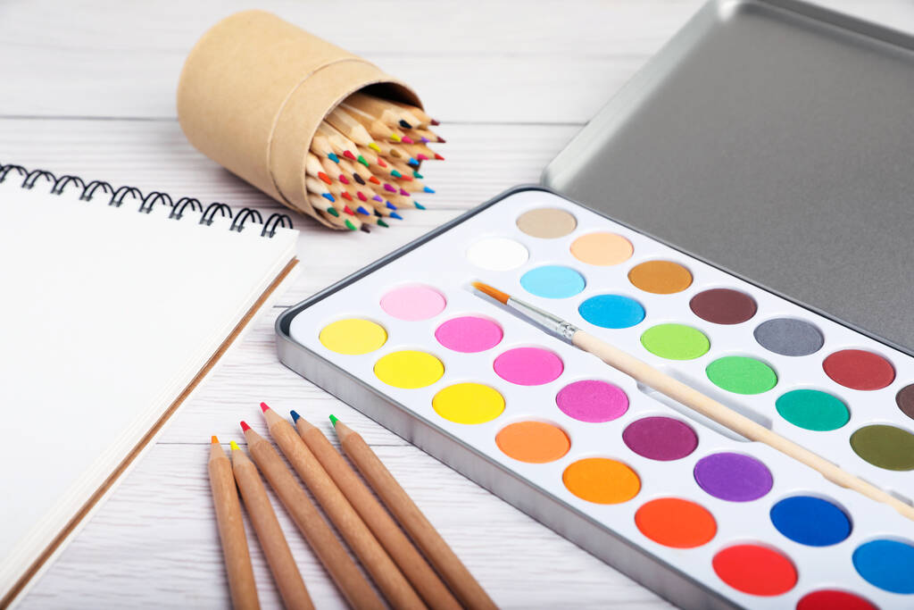 Aquarelpalet met penseel, kleurrijke potloden en notitieboekje op witte houten tafel, close-up - Foto, afbeelding