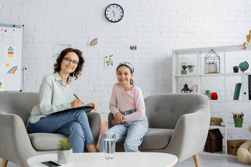 Vrolijke psycholoog schrijven op klembord en kijken naar camera in de buurt van tiener meisje in consulting kamer  - Foto, afbeelding