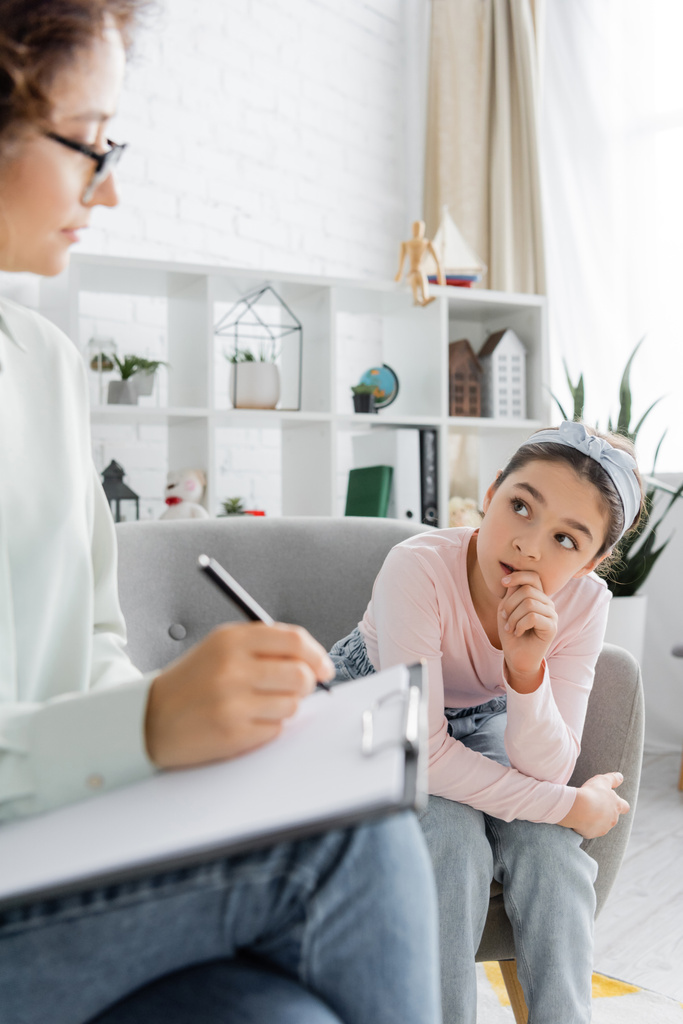 tiener tiener meisje kijken wazig psycholoog schrijven op klembord in consulting kamer  - Foto, afbeelding