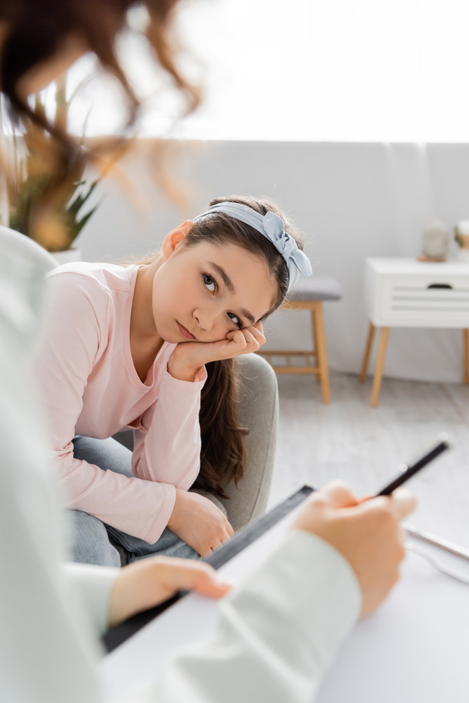 Sad preteen child looking at blurred psychologist with clipboard in consulting room  - Photo, Image
