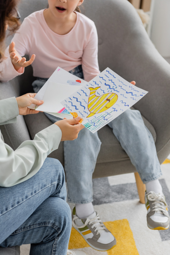 Cropped view of psychologist holding drawings while preteen patient talking in consulting room  - Photo, Image