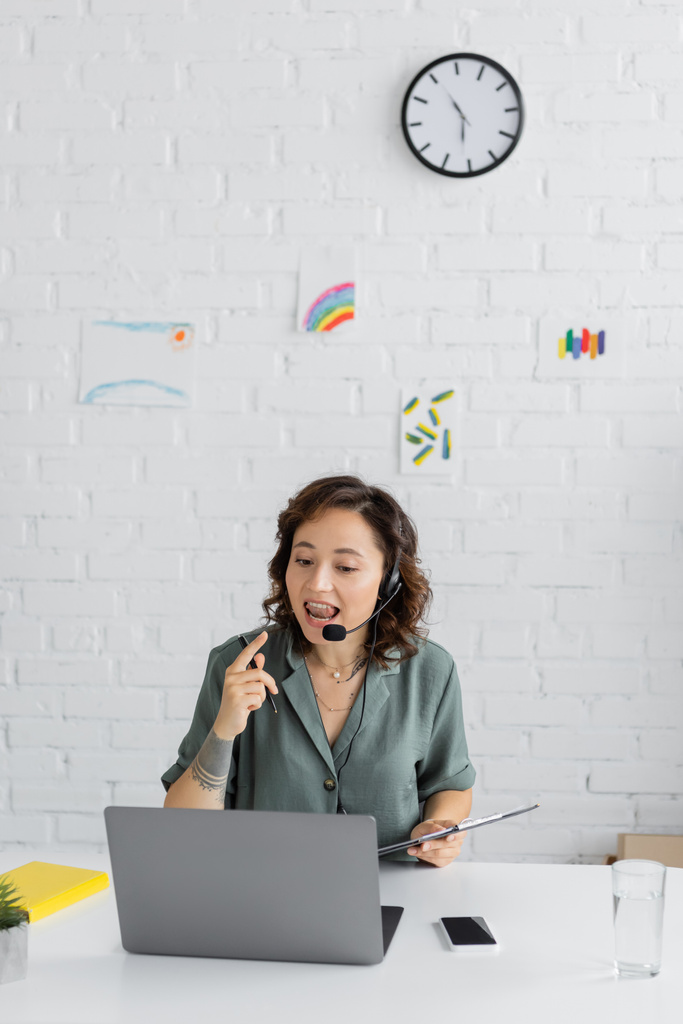 Spraaktherapeut in headset praten en houden klembord tijdens videogesprek op laptop in consulting room  - Foto, afbeelding