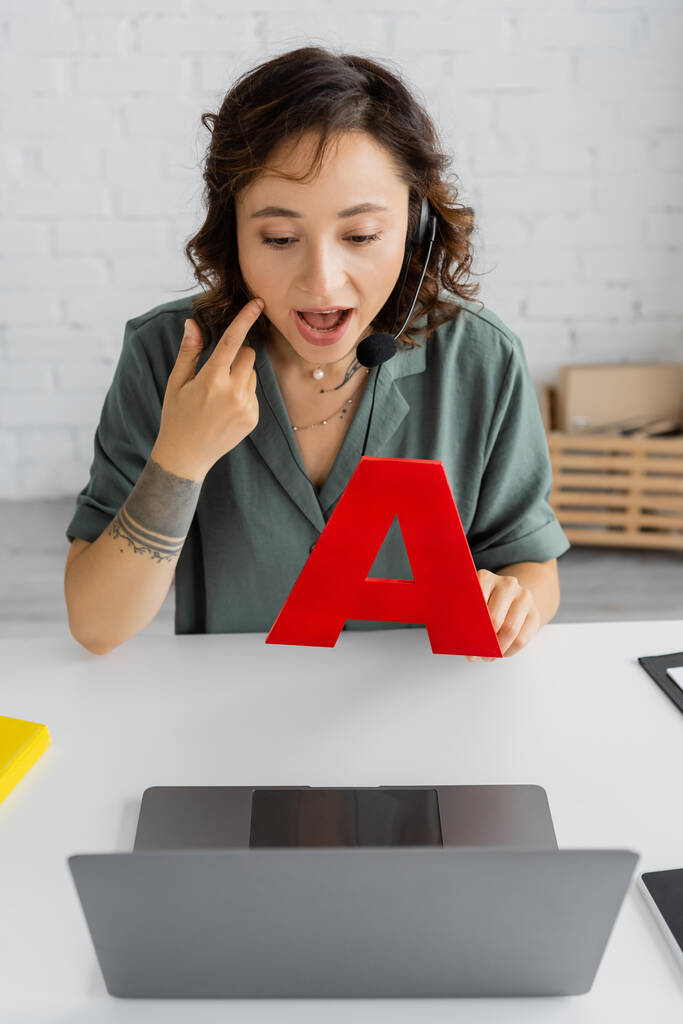 Logopeda dotykająca policzka i trzymająca list podczas lekcji video na laptopie w pokoju konsultacyjnym  - Zdjęcie, obraz