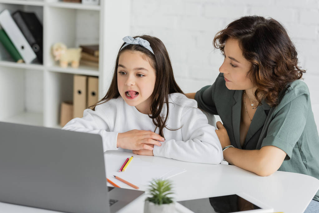 Genitore seduto vicino a figlia con logopedia lezione online vicino ai dispositivi a casa  - Foto, immagini