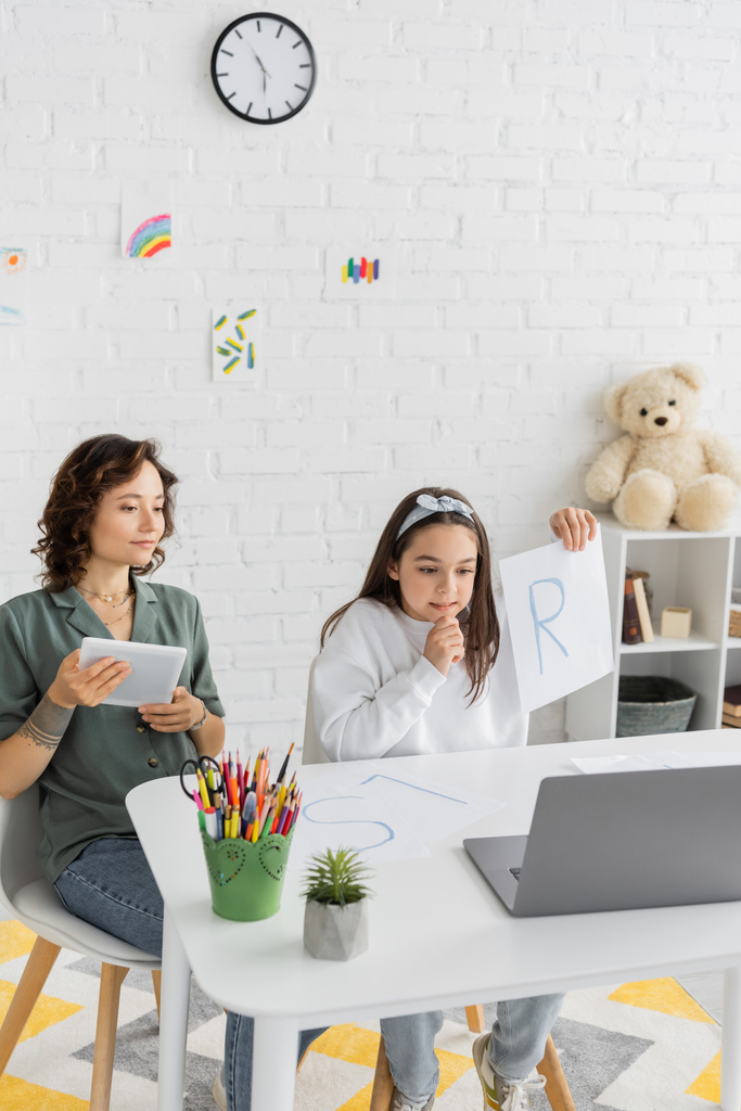 Позитивна дитина тримає папір з листом і розмовляє під час мовної терапії онлайн урок біля мами вдома
  - Фото, зображення