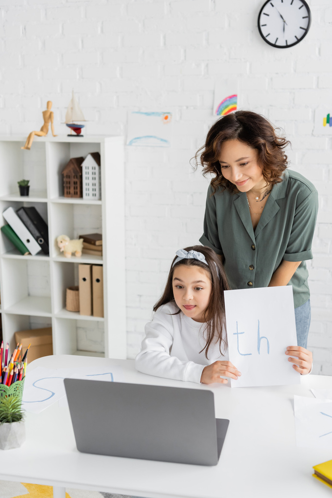 Улыбающаяся мать, стоящая рядом с дочерью, держит бумагу с письмами во время онлайн-урока логопедии дома  - Фото, изображение