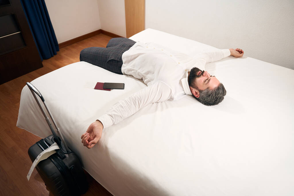 Mosolygó elégedett elegáns férfi feküdt az ágyon a jól megvilágított szállodai szobában - Fotó, kép