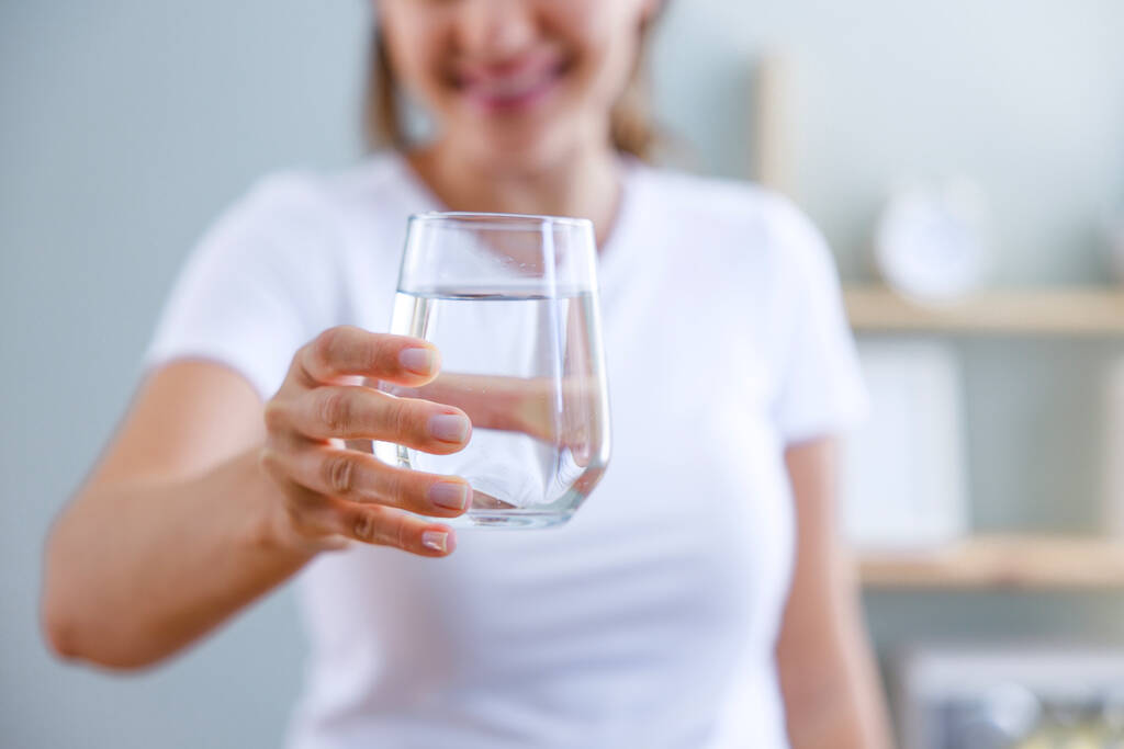 Une jeune femme souriante montrant un grand verre d'eau dans sa main. La mise au point se fait dans un grand verre d'eau. Avantages pour la santé de boire suffisamment d'eau concepts.  - Photo, image