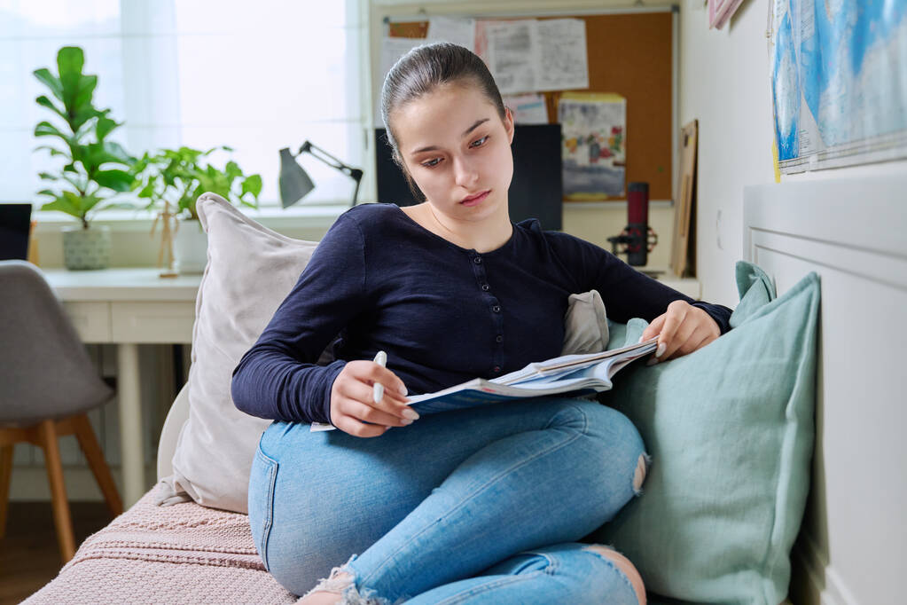 Teinityttö opiskelee kotona, lukee oppikirjaa, kirjoittaa muistikirjaan, istuu sohvalla kotona. Nuoruus, lukiolainen, koulutustiedon käsite - Valokuva, kuva