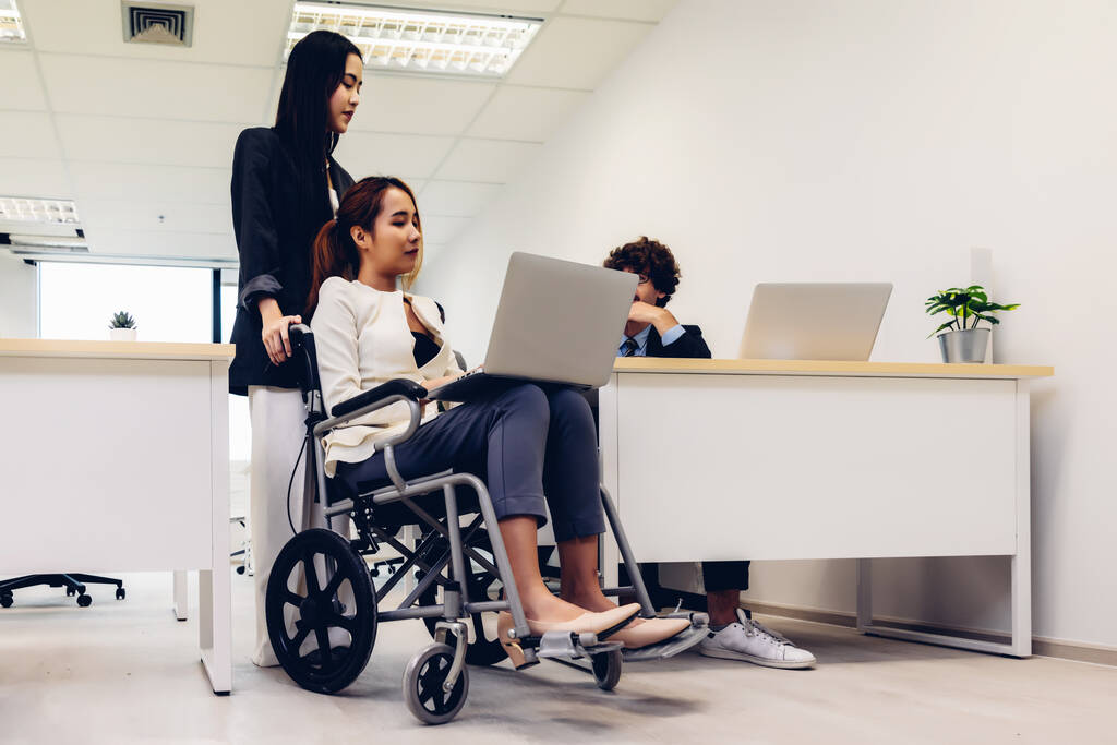 明るいオフィスの車椅子のオフィスの労働者と女性。彼らはチームワークを示しています。オフィスのカメラで笑顔で車椅子の若い女性と多様なビジネスチームの肖像. - 写真・画像