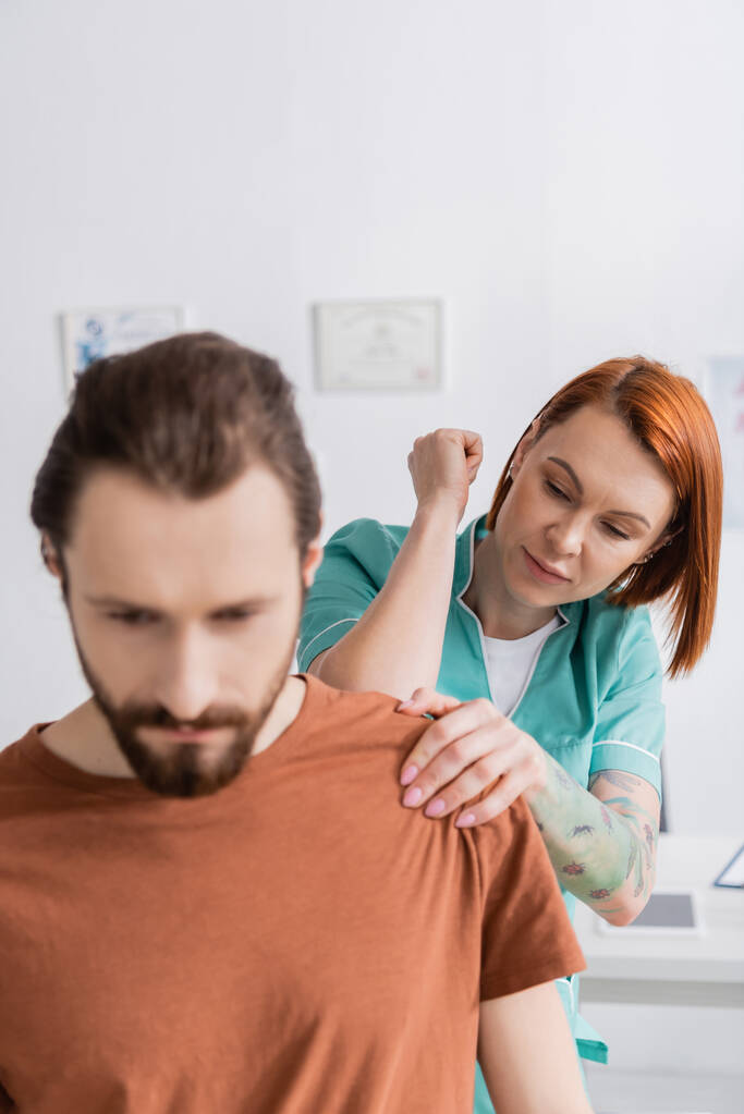 fyzioterapeut vyšetřující poraněné rameno vousatého muže v rehabilitačním centru - Fotografie, Obrázek