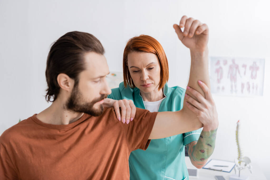 pelirroja fisioterapeuta flexión brazo y tocando hombro de hombre lesionado en la sala de consulta - Foto, Imagen