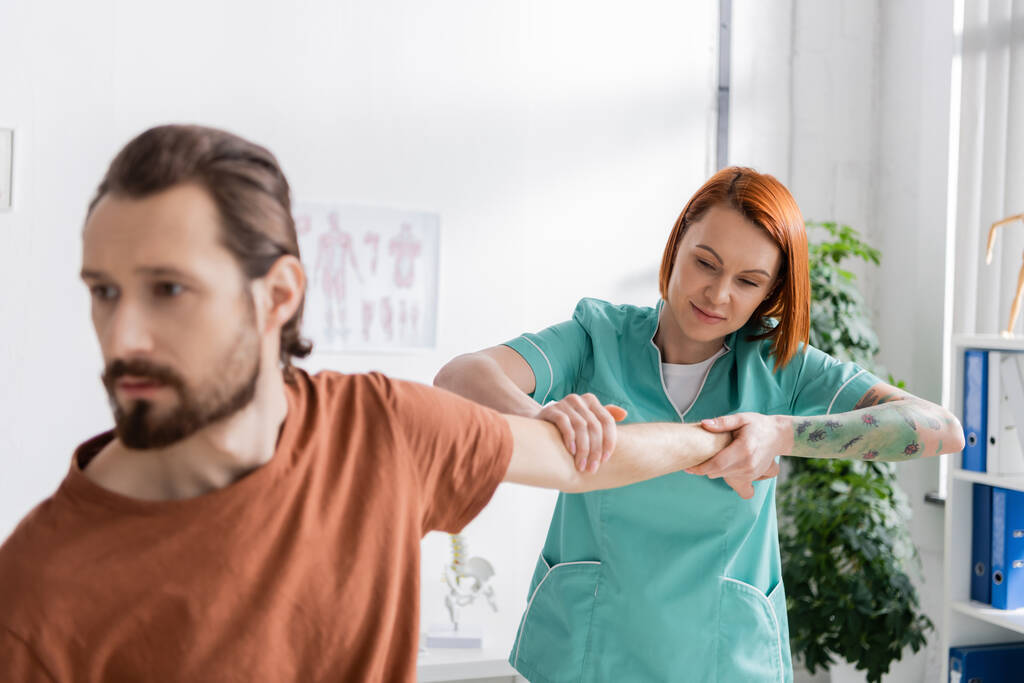 redhead physiotherapist stretching arm of blurred man during diagnostics in rehab center - Photo, Image