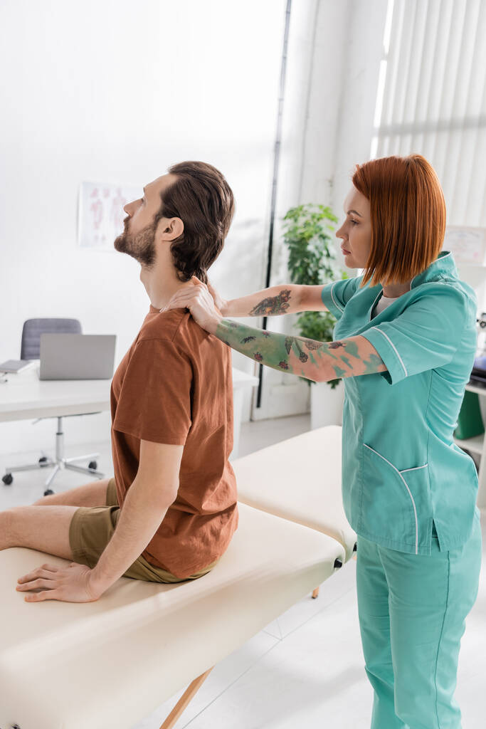 vue latérale d'un ostéopathe examinant le dos d'un homme barbu assis sur une table de massage dans une salle de consultation - Photo, image