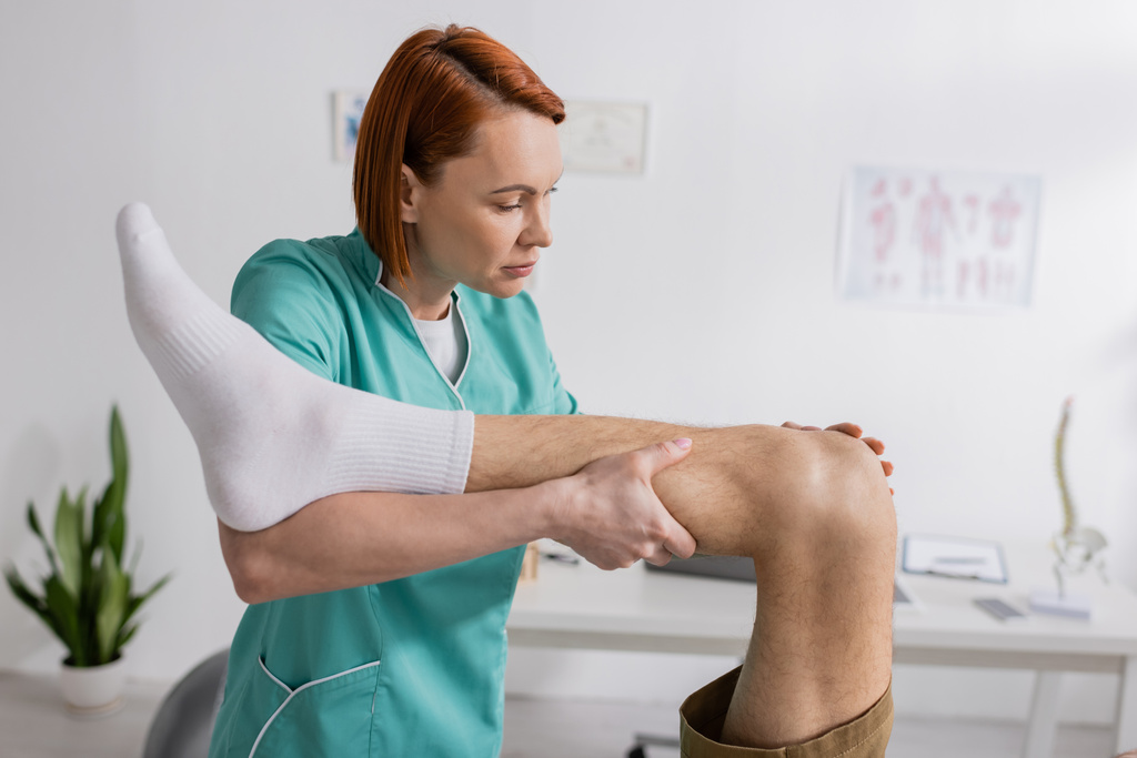 Rotschopf-Handtherapeut arbeitet mit verletztem Bein von Mann in Reha-Zentrum - Foto, Bild