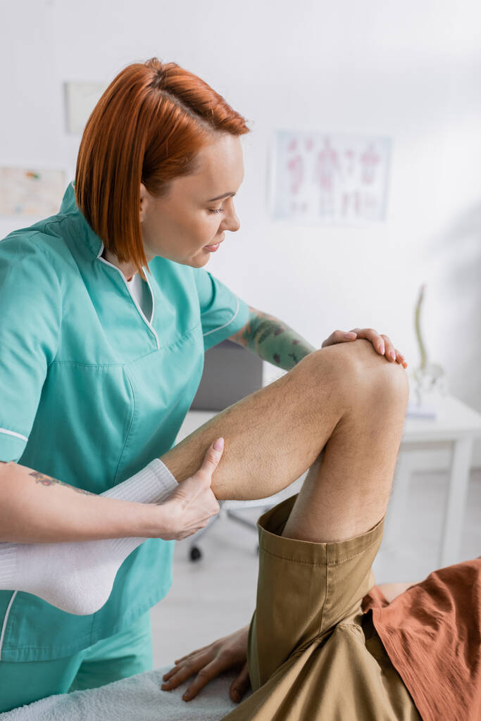 roodharige fysiotherapeut buigt been van gewonde man tijdens revalidatietherapie in spreekkamer - Foto, afbeelding