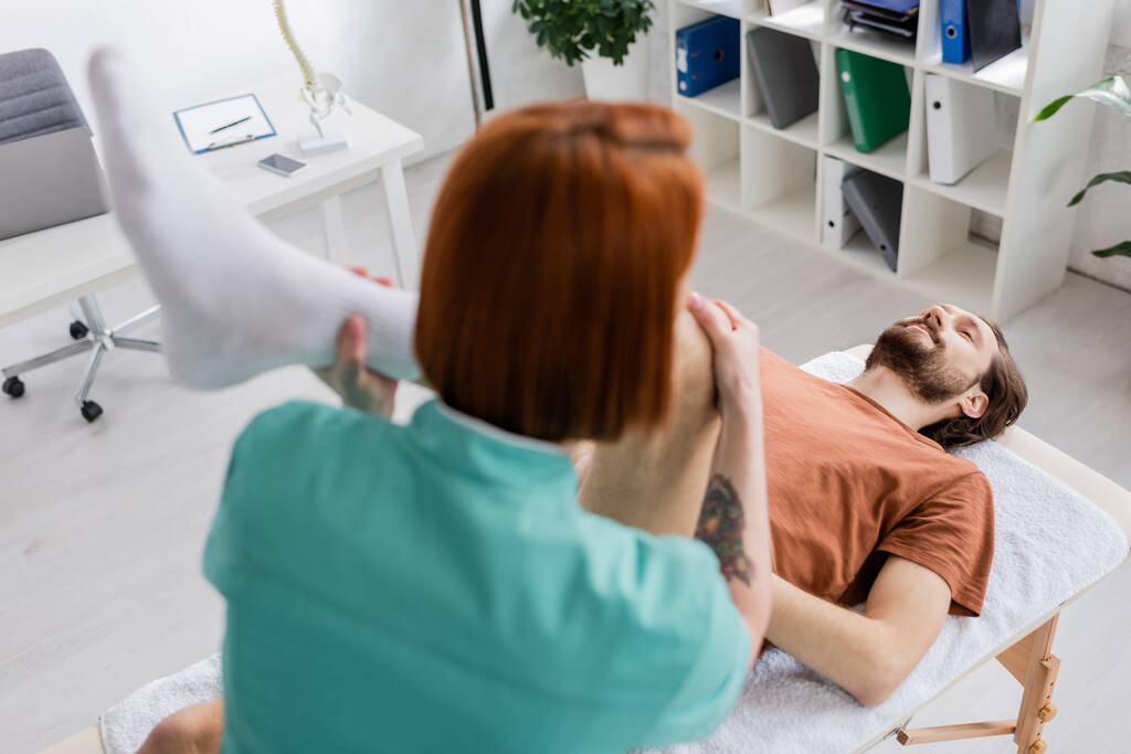wazig manuele therapeut buigen gewond been van de mens terwijl het maken van pijnstillende massage in revalidatiecentrum - Foto, afbeelding