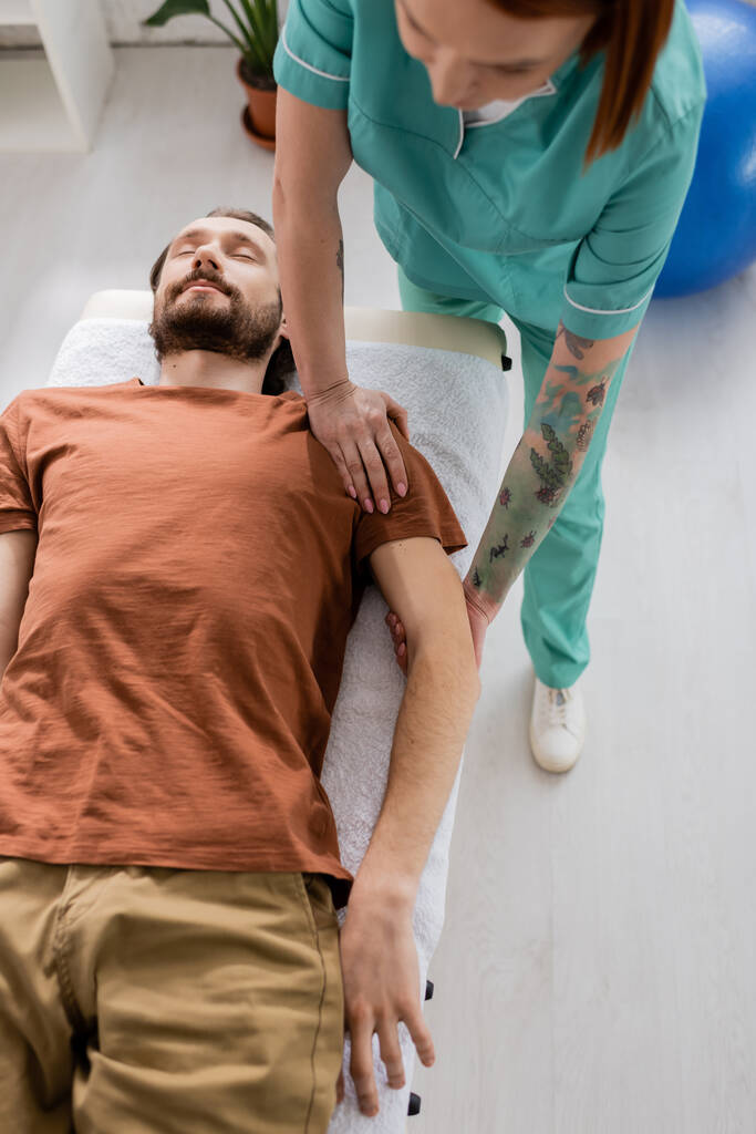 vista de ángulo alto del hombre barbudo acostado cerca de fisioterapeuta haciendo masaje de brazo y hombro en el centro de recuperación - Foto, Imagen