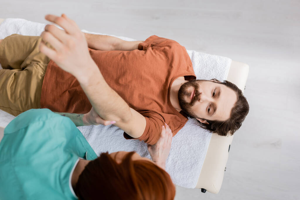 вид зверху розмитого мануального терапевта масажу пораненої руки бородатого чоловіка в реабілітаційному центрі
 - Фото, зображення
