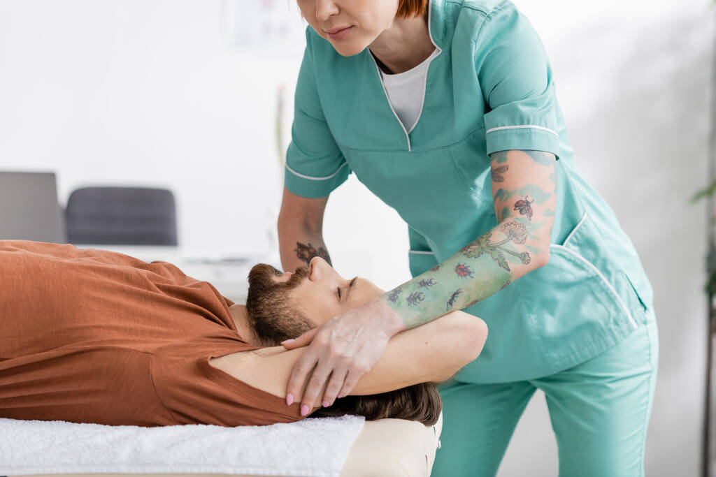татуйований фізіотерапевт масажує болючу руку бородатого чоловіка під час лікування в реабілітаційному центрі
 - Фото, зображення