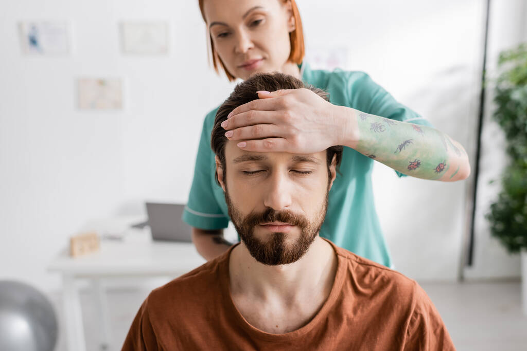 verschwommener Physiotherapeut berührt Stirn eines bärtigen Mannes mit geschlossenen Augen bei Diagnostik im Sprechzimmer - Foto, Bild