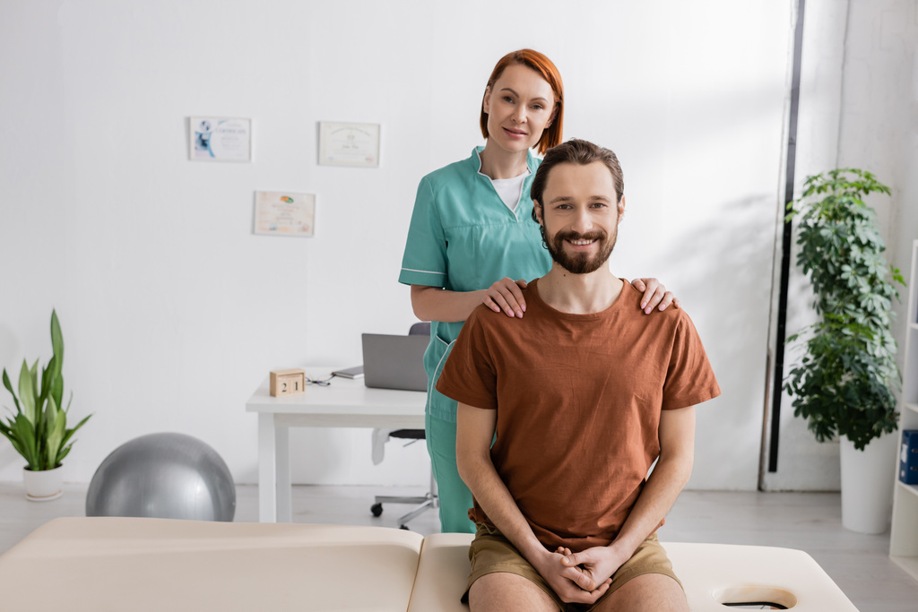 счастливый физиотерапевт трогает плечи бородатого мужчины, сидящего на массажном столе в кабинете - Фото, изображение