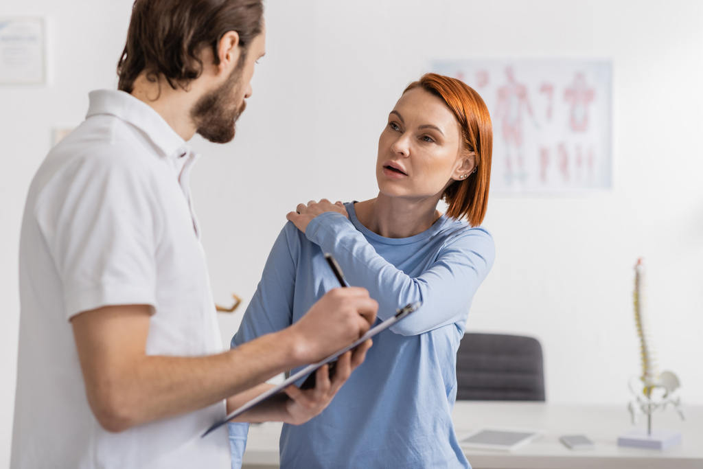 donna rossa con spalla ferita che parla con fisioterapista scrivendo diagnosi negli appunti in ospedale - Foto, immagini