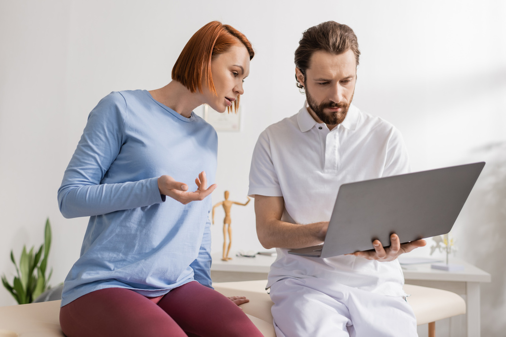 physiothérapeute barbu à l'aide d'un ordinateur portable près rousse femme pointant avec la main dans la salle de consultation - Photo, image