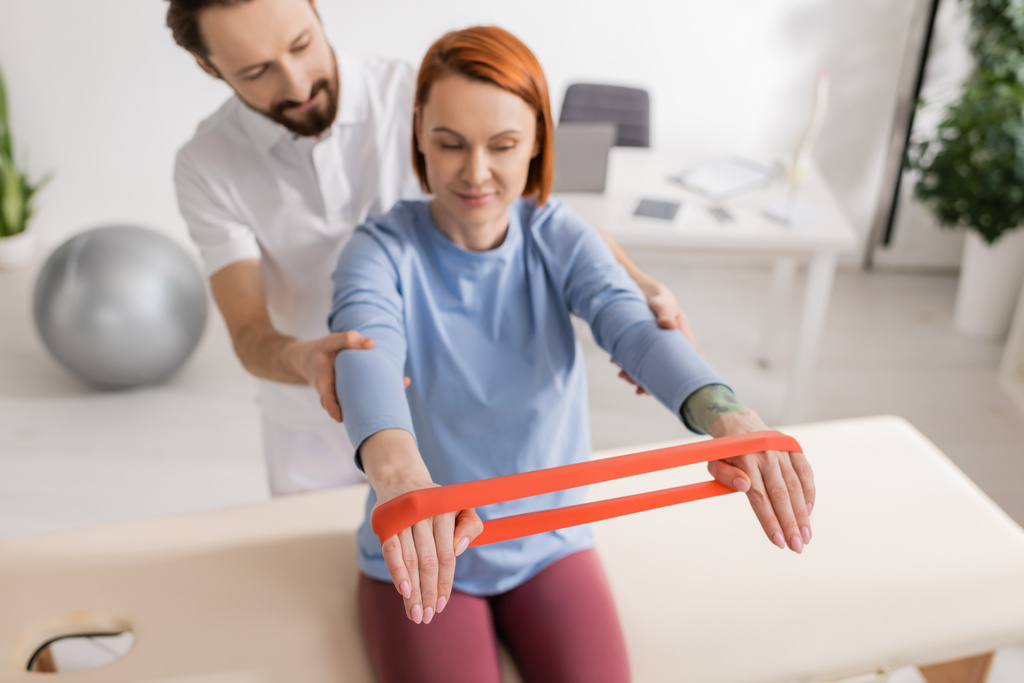 physiotherapist supporting smiling woman working out with resistance band on massage table in clinic - Photo, Image