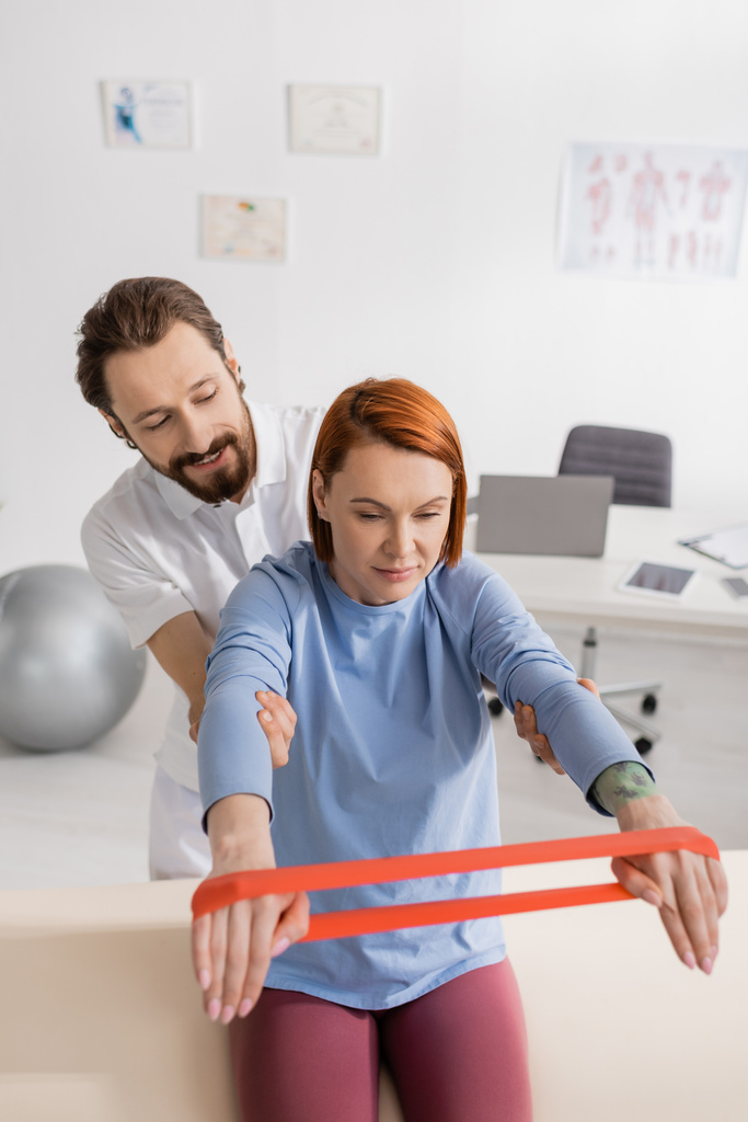 smiling physiotherapist supporting redhead woman training arms with resistance band in rehab center - Photo, Image