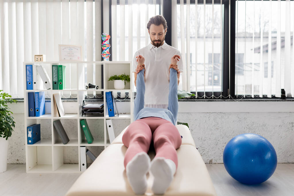fisioterapeuta barbudo estirar los brazos dolorosos de la mujer acostada en la mesa de masaje en el centro de rehabilitación moderno - Foto, Imagen